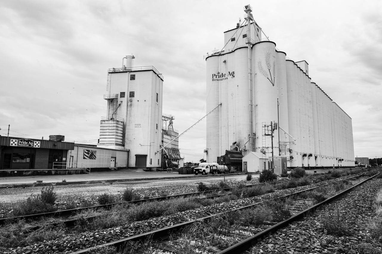 Black and white photo of grain silos by railway tracks under cloudy skies.